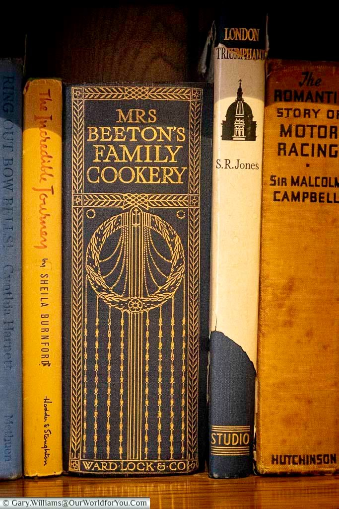 The image shows a close-up of several books standing upright on a shelf. The most prominent book is "Mrs. Beeton's Family Cookery," featuring a dark blue cover with ornate gold lettering and a decorative illustration.