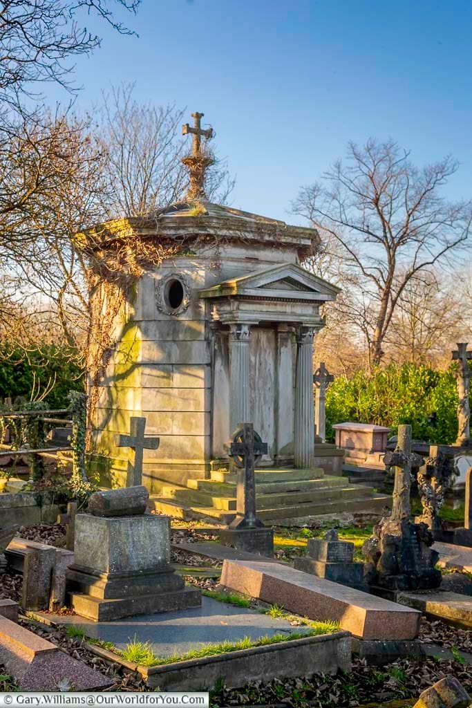 The image shows a Victorian-era mausoleum with a prominent cross on top, situated amongst numerous other gravestones and crosses in West Norwood Cemetery, London. Bare winter trees cast long shadows across the scene under a clear, pale blue sky.