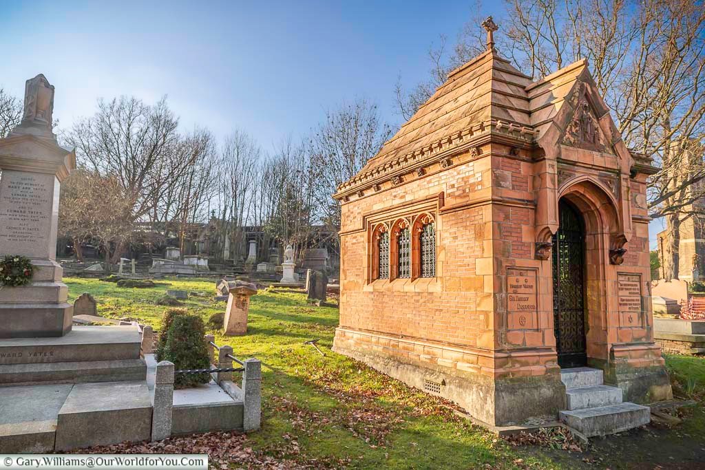 The image shows the striking terracotta mausoleum of Sir Henry Doulton in West Norwood Cemetery, London, bathed in warm sunlight under a clear blue sky. The ornate Gothic-style structure stands prominently amongst other weathered gravestones and monuments in the historic cemetery.