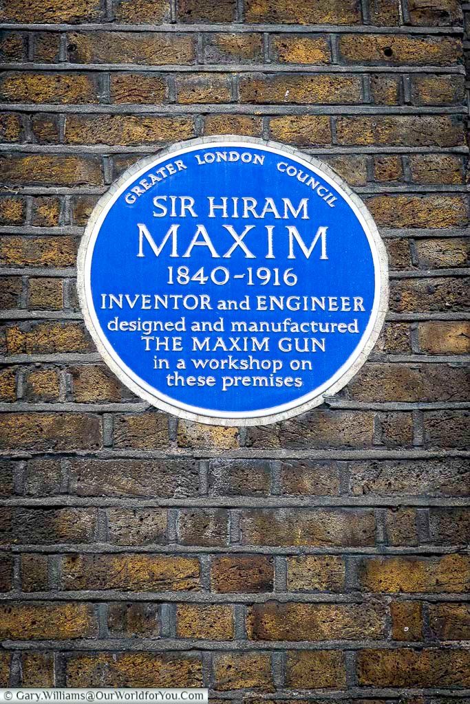 The image shows a blue Greater London Council plaque affixed to a weathered brick wall. The plaque commemorates Sir Hiram Maxim (1840-1916), an inventor and engineer who designed and manufactured the Maxim gun in a workshop on these premises.
