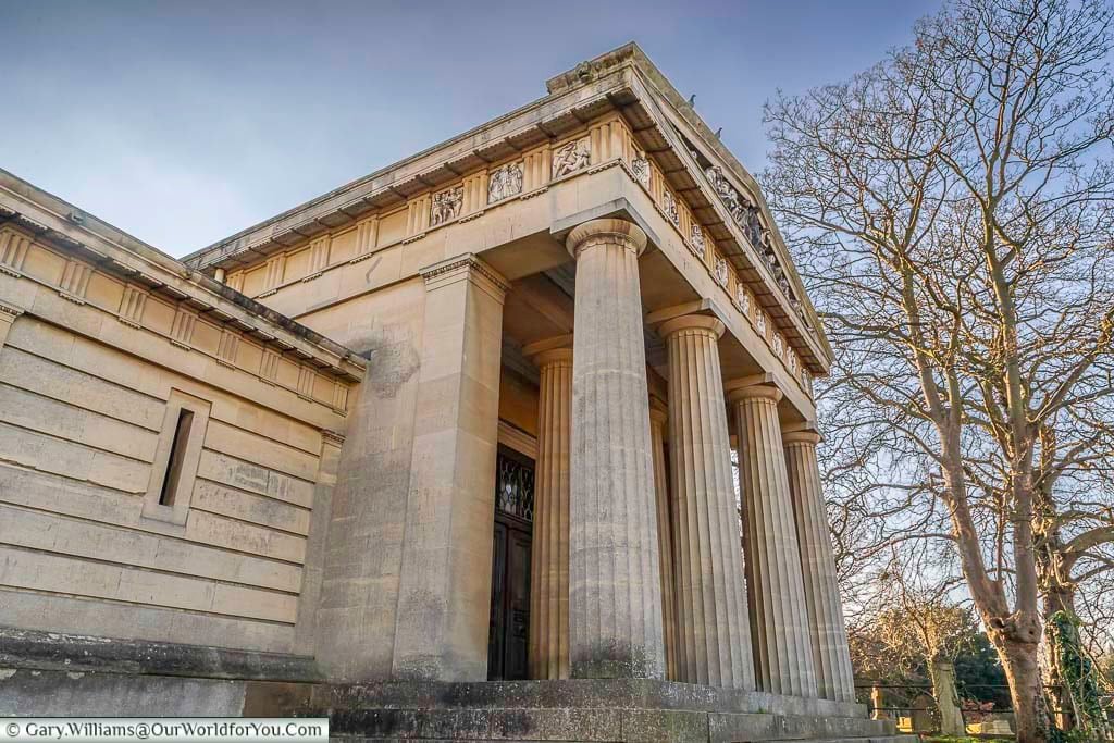 The image shows the grand, neo-classical entrance to a building within West Norwood Cemetery, London, featuring large stone columns and intricate architectural details. The structure stands under a soft, slightly overcast sky, with bare winter trees visible to the right.