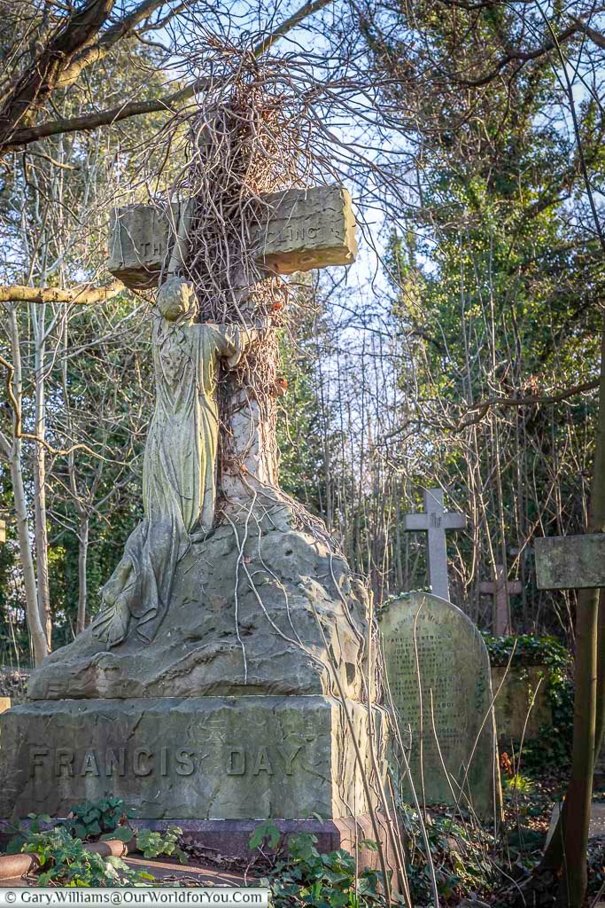 The image shows the weathered and vine-covered grave of Francis Day in West Norwood Cemetery, London, featuring a large stone cross with a sculpted figure embracing it. Sunlight filters through the surrounding trees, illuminating the memorial and the serene, overgrown setting of this historic cemetery.