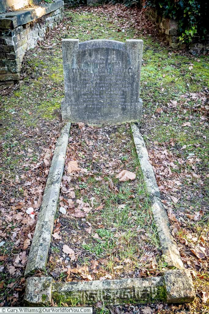 The image shows a weathered gravestone with surrounding stone borders in West Norwood Cemetery, London, amidst fallen leaves and patches of green moss. The inscription on the headstone is worn and partially obscured, hinting at the passage of time in this historic burial ground.