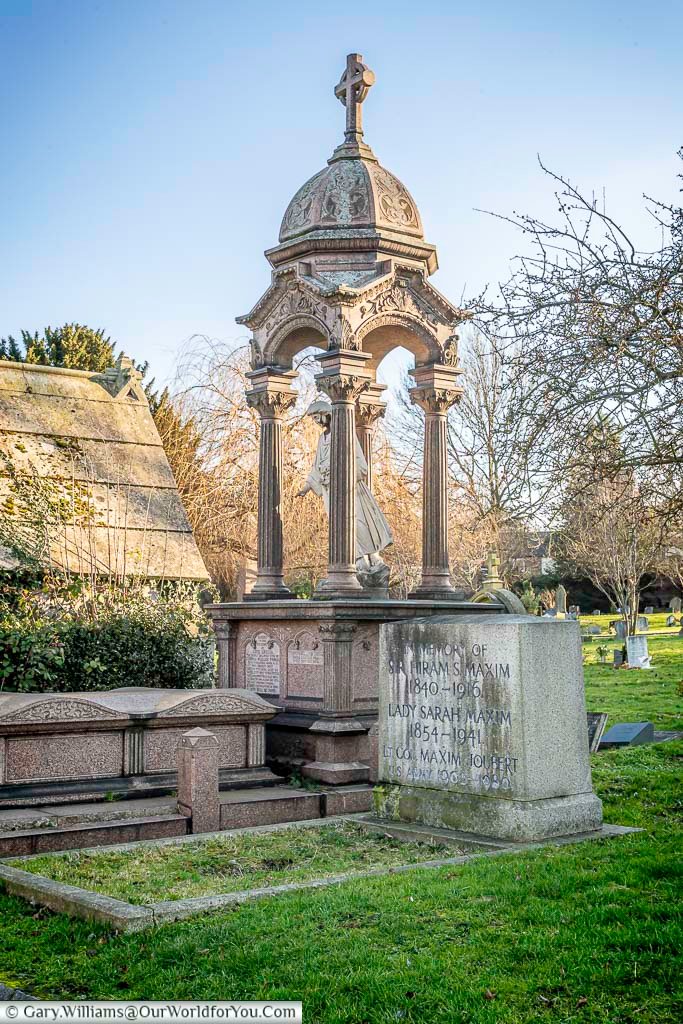 The image shows the elaborate memorial to Sir Hiram Maxim and his family in West Norwood Cemetery, London, featuring an ornate stone canopy supported by columns and topped with a cross. A statue of a winged figure stands beneath the canopy, and inscribed stone blocks detail the names and dates of the deceased.