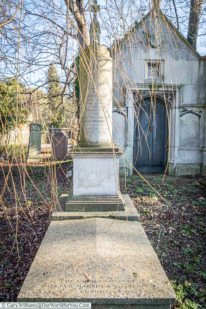 The image shows a tall, cylindrical gravestone with inscriptions, situated in West Norwood Cemetery, London, surrounded by fallen leaves and the bare branches of trees under a bright sky. In the background stands a weathered stone mausoleum with a dark, arched doorway, typical of the Victorian architecture found in this historic cemetery.
