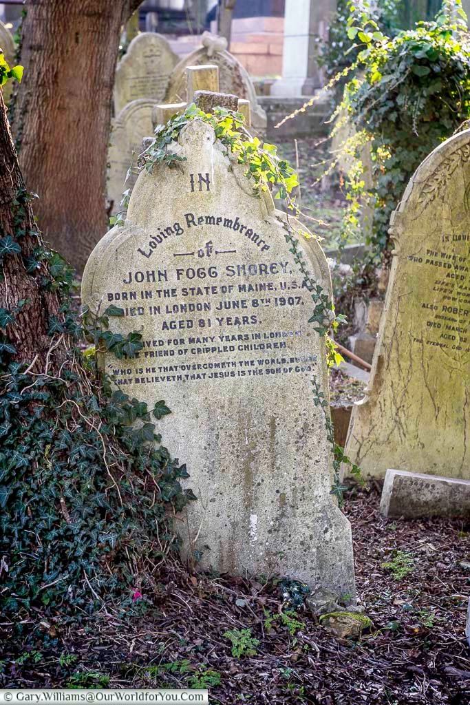 The image shows a weathered gravestone in West Norwood Cemetery, London, partially covered in ivy. The inscription reads "In Loving Remembrance of John Fogg Shorey, born in the State of Maine, U.S.A., died in London June 6th 1907, aged 81 years."