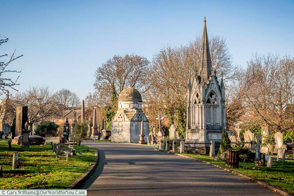 The image shows a paved pathway winding through West Norwood Cemetery in London on a sunny day. Elaborate Victorian-era tombs and monuments of various styles, including a tall Gothic spire and a domed structure, line both sides of the path, set amongst green grass and bare winter trees under a clear blue sky.