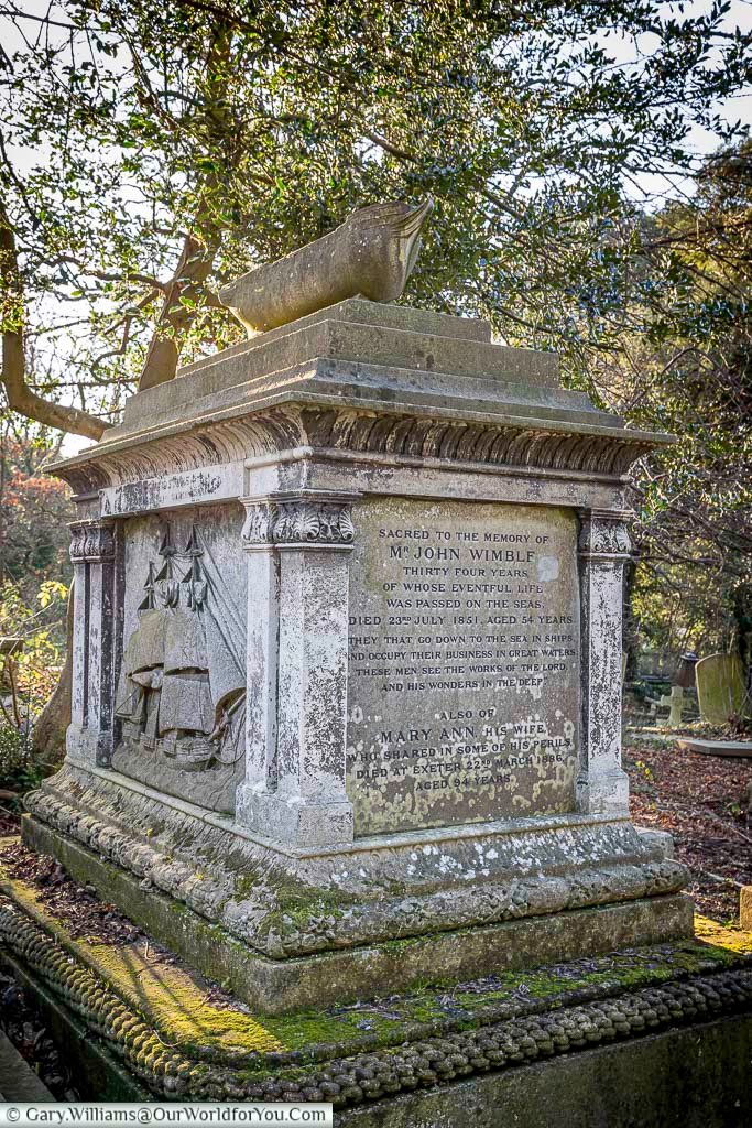 The image shows a weathered stone tomb adorned with carvings and inscriptions, topped with a sculpture of a ship. The inscription commemorates John Wimble, noting his 34 years at sea, and his wife Mary Ann, who shared in some of his perils.