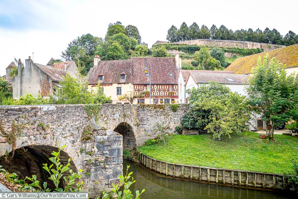 A picturesque view captures a stone arched bridge in Semur-en-Auxois, France, spanning a calm waterway. Historic buildings with terracotta roofs and varying facades line the waterway beyond the bridge, nestled against a backdrop of lush green trees and a stone wall along the upper edge. The scene exudes a tranquil and timeless charm under a soft, overcast sky.