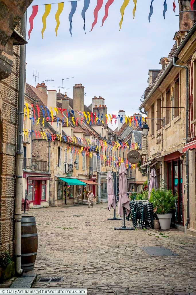 A slightly low-angle shot captures a charming, narrow cobblestone street winding gently through the medieval town of Semur-en-Auxois, France. Colorful triangular pennants in red, yellow, blue, and white stretch across the street overhead, creating a festive atmosphere. Buildings line both sides of the street, showcasing a variety of architectural styles with light-colored stone facades, some featuring balconies with wrought-iron railings and vibrant awnings in red and green.