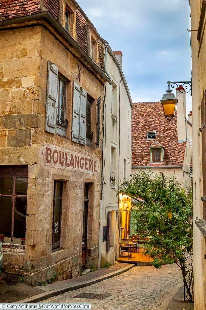 This street-level shot captures a charming, narrow cobblestone street in Semur-en-Auxois, France, lined with historic buildings. On the left, a weathered stone building features faded red lettering that reads "BOULANGERIE" above its storefront with dark windows and grey shutters. The street curves gently, revealing a warmly lit outdoor seating area of a restaurant further down, creating a cozy and inviting atmosphere under a dusky sky.