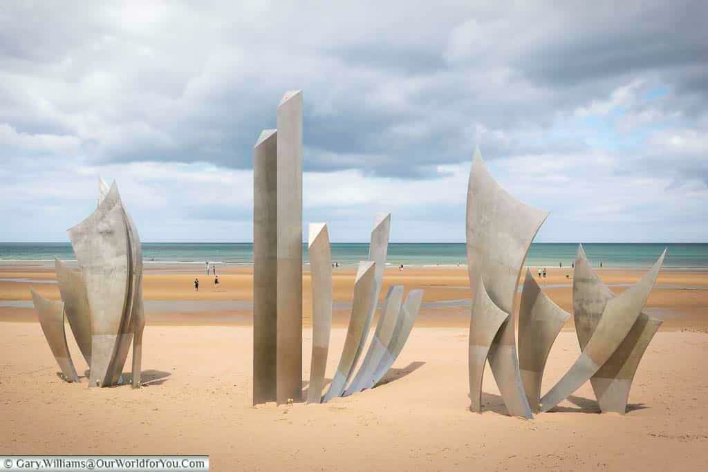 The striking "Les Braves" monument, a series of sharp, angled steel sculptures, stands prominently on the sandy expanse of Omaha Beach. These powerful figures face the sea, a poignant reminder of the courage and sacrifice of the soldiers who landed here during D-Day under a dramatic, partly cloudy sky.