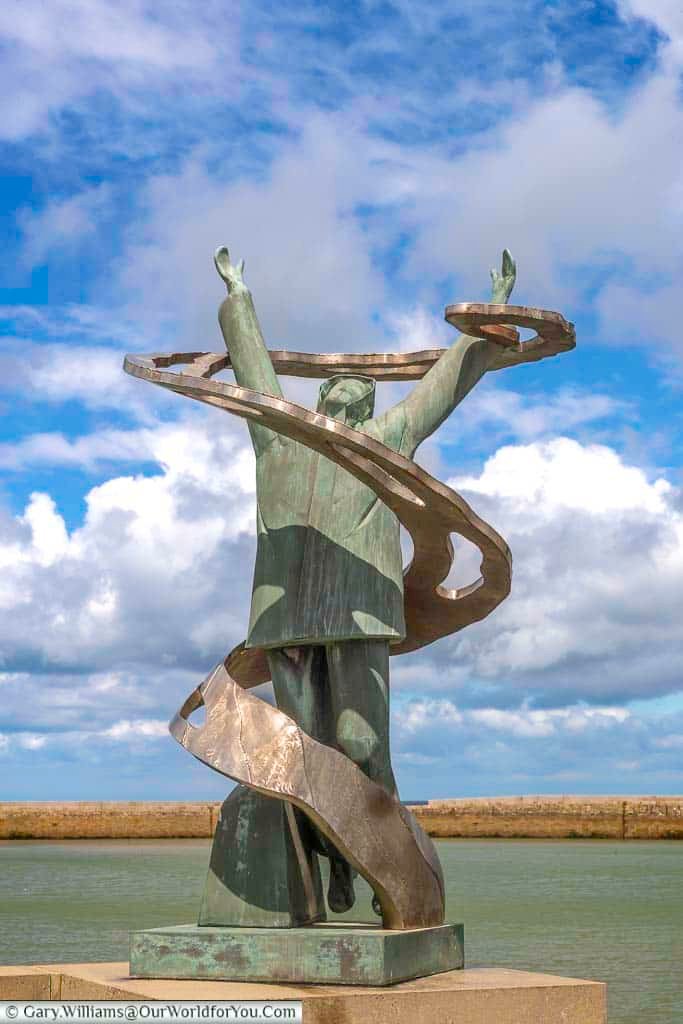 The image shows a bronze sculpture of a man with his arms raised, surrounded by swirling metal bands, set against a backdrop of a cloudy blue sky and a harbor. This monument commemorates sailors who perished at sea in Port-en-Bessin, located in the Calvados region of Normandy.