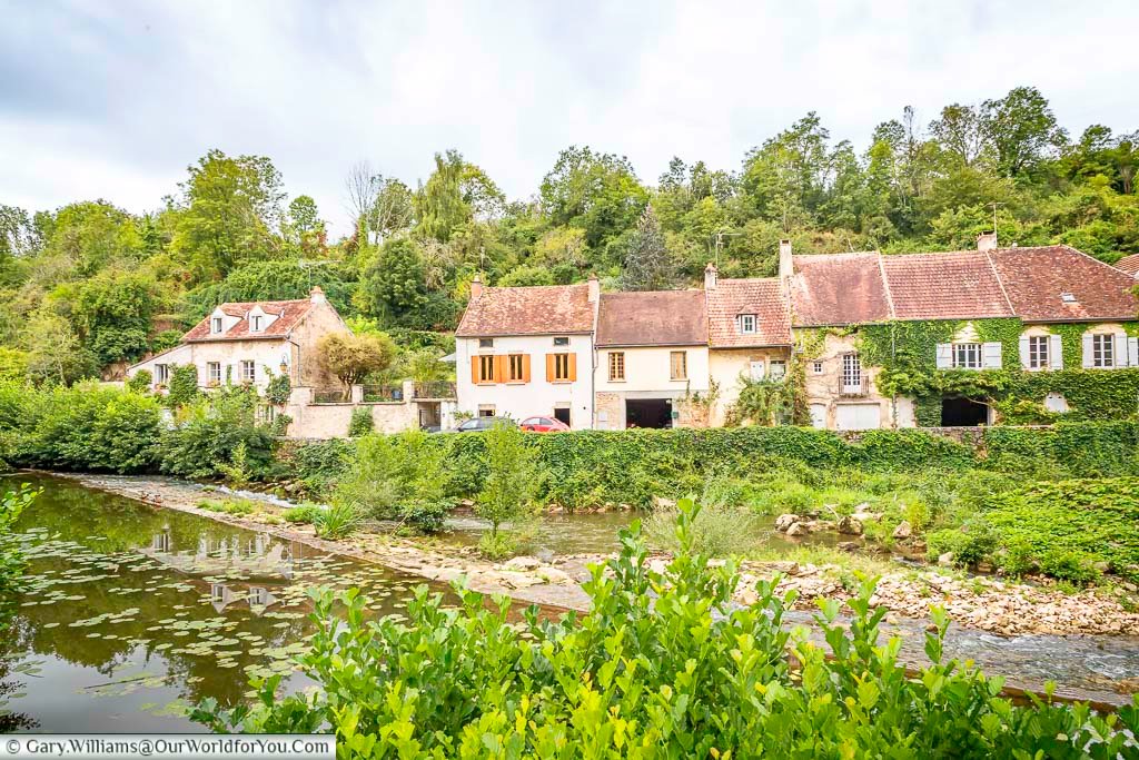 A serene waterside view showcases a row of charming houses in Semur-en-Auxois, France, nestled along a gently flowing river. The houses feature terracotta-tiled roofs and a mix of light-colored facades, with one partially covered in lush green ivy. Verdant foliage lines the riverbanks and the hillside rising behind the houses, creating a peaceful and picturesque scene under a soft, overcast sky.