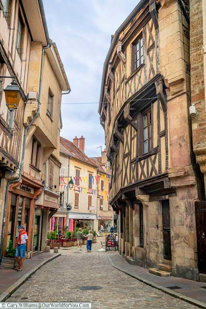 A street-level shot looks down a narrow, winding cobblestone street in Semur-en-Auxois, France, framed by charming medieval buildings. The buildings on either side feature a mix of light-colored stone and distinctive half-timbered facades with dark wooden beams. Colorful triangular pennants are strung overhead, adding a festive touch to the quaint street scene where a few pedestrians stroll along.