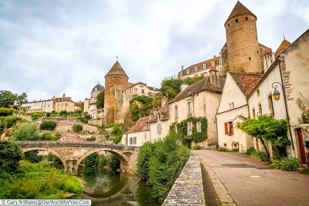 A picturesque view captures the medieval town of Semur-en-Auxois, France, perched along the banks of a calm river. An arched stone bridge, likely the Pont Pinard, spans the water, reflecting its structure and the surrounding buildings in the gentle current. The town's ancient stone walls and a prominent round tower rise above the riverbank, nestled amongst charming buildings and lush greenery under an overcast sky.