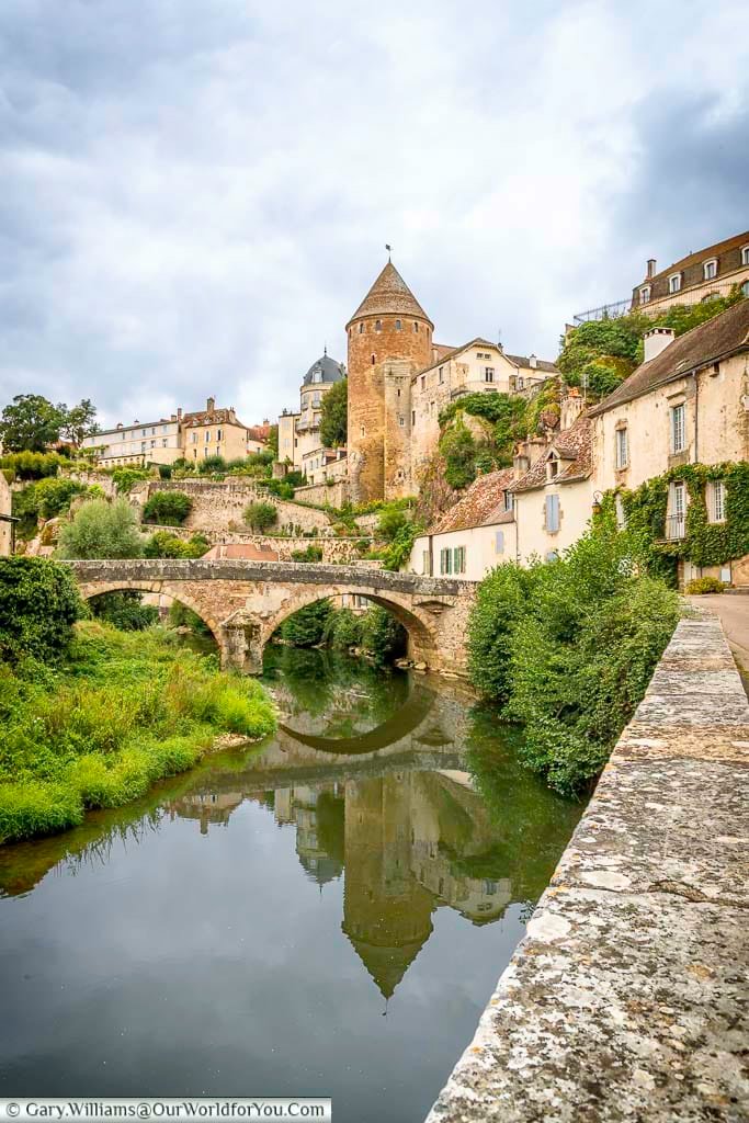 A scenic view captures the medieval town of Semur-en-Auxois, France, nestled along a tranquil river spanned by an arched stone bridge. The town's ancient stone buildings, including prominent cylindrical towers with conical roofs, rise along the hillside, their reflections visible in the calm water below. Lush greenery lines the riverbanks and weaves through the architecture, creating a picturesque blend of nature and history under an overcast sky.