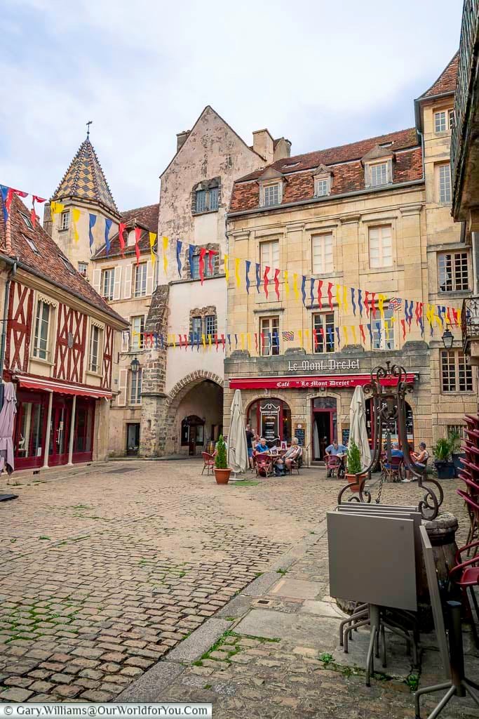 This eye-level shot captures a lively square in Semur-en-Auxois, France, adorned with colorful triangular banners strung between the historic buildings. The cobblestone square features outdoor seating areas with patrons enjoying themselves under red awnings of a café named "Le Mont Drejet." Architectural details include a turret with a patterned roof on the left and various light-colored stone facades with shuttered windows, creating a charming medieval ambiance.