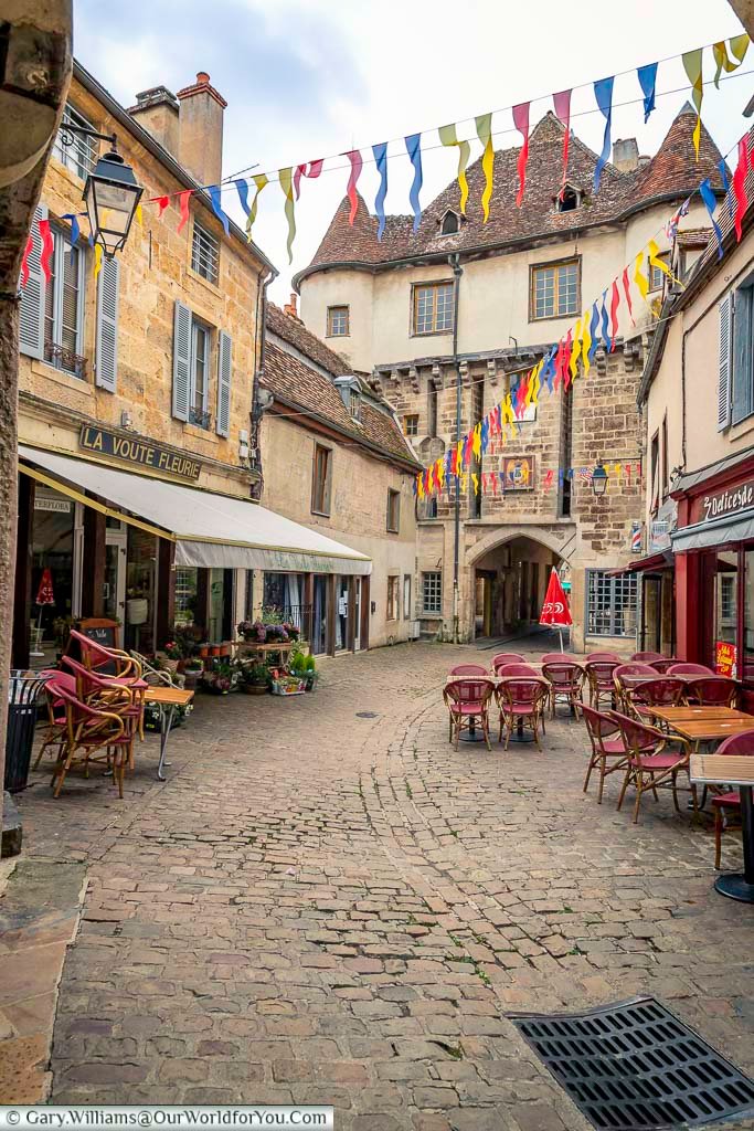A charming square in Semur-en-Auxois, France, is decorated with colorful pennants strung above the aged cobblestone pavement. Shops with outdoor seating and vibrant floral displays line the square, leading towards an arched stone gateway in the background.