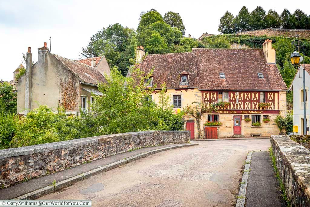 A charming street scene in Semur-en-Auxois, France, unfolds with a stone bridge leading towards historic buildings nestled against a lush, tree-covered hillside. The building on the right features distinctive half-timbered upper levels with red cross bracing and a sign reading "Maison Rose 'Les Minimes'." The weathered stone of the bridge and the varied textures of the buildings evoke the town's rich history under a soft, overcast light.