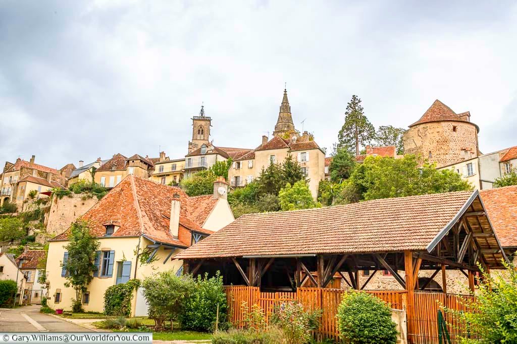 This slightly low-angle shot captures a picturesque view of Semur-en-Auxois, France, with its cluster of historic buildings nestled on a hillside under a cloudy sky. In the foreground, terracotta-tiled roofs of lower buildings and lush greenery lead the eye up to the town's skyline, punctuated by the spires of the Collegiate Church of Notre-Dame and the cylindrical Tour de l'Orle d'Or. The varying architectural styles and the way the town cascades down the slope create a charming and timeless scene.