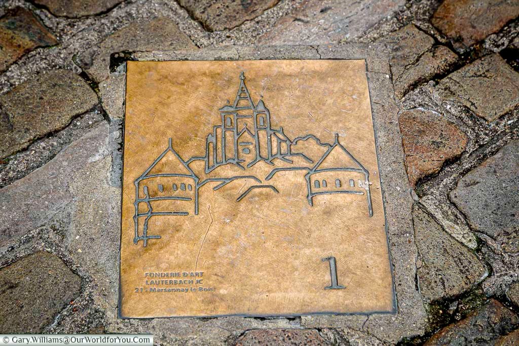 This close-up shot focuses on a square, bronze plaque embedded in the cobblestone pavement of Semur-en-Auxois, France. The plaque features a stylized line drawing of the town's distinctive architecture, including its prominent towers and buildings clustered on a hill. Inscribed on the lower left are the words "FONDERIE D'ART / LAUTERBACH JC / 21210 MARSANNAY le BOIS," and a large numeral "1" is visible on the lower right.