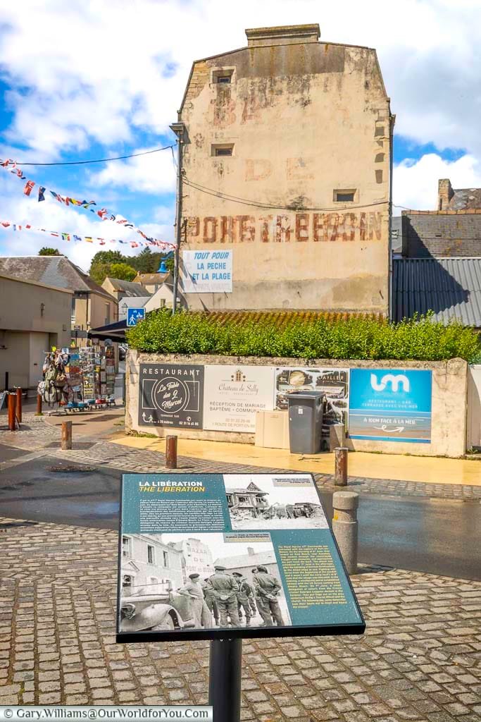 The image shows a street corner in a historic town of Port-en-Bessin, Normandy, featuring weathered stone buildings with faded signage and colorful bunting strung across the street. In the foreground, an information board titled "LA LIBÉRATION DE PORT-EN-BESSIN" displays historical photographs and text related to the town's liberation during World War II.