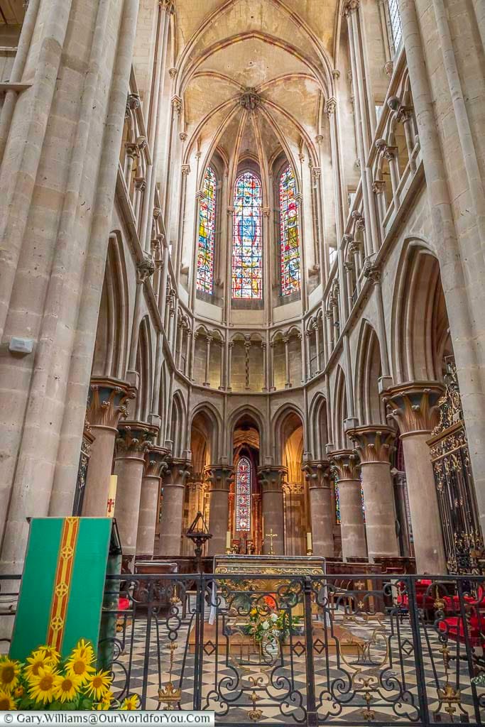 This interior shot captures the grandeur of the Collegiate Church of Notre-Dame in Semur-en-Auxois, France, showcasing its soaring Gothic architecture. Tall, slender columns support pointed arches and ribbed vaults that reach towards the high ceiling. Stained glass windows flood the apse with colorful light, illuminating the altar area and the intricate details of the stone carvings.