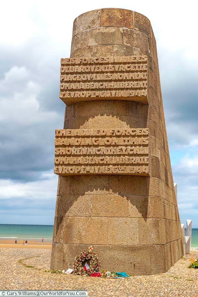 A tall, cylindrical stone monument stands prominently on the sandy expanse of Omaha Beach, under an overcast sky. Inscribed text in both French and English commemorates the Allied forces who landed on this shore on D-Day, June 6th, 1944, to liberate Europe, and a wreath lies at its base as a tribute.