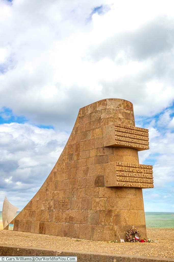A unique, sloped stone monument stands on the sandy shores of Omaha Beach under a cloudy sky. Inscriptions on the monument commemorate the Allied forces who liberated Europe at Omaha Beach.