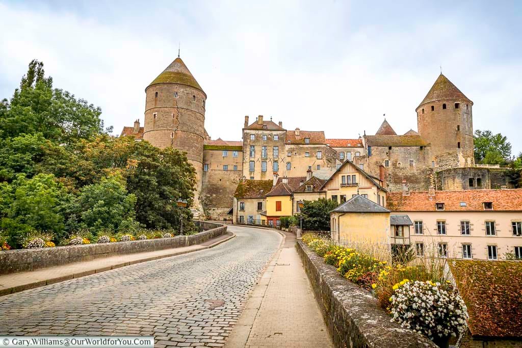 A wide, slightly low-angle shot captures a road leading into the medieval town of Semur-en-Auxois, France, showcasing its impressive stone fortifications and historic buildings. Two prominent cylindrical towers with conical roofs stand guard over the town, which is nestled on a rocky outcrop overlooking the surrounding landscape. Lush greenery and colorful flowers line the sides of the road, adding a touch of vibrancy to the ancient architecture under a cloudy sky.
