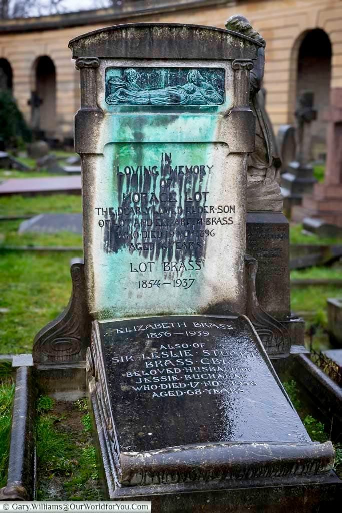 This close-up shot shows a weathered gravestone in Brompton Cemetery, United Kingdom, with a bronze relief panel at the top depicting figures. The stone bears inscriptions "IN LOVING MEMORY OF HORACE LOT BRASS" and details for other family members.