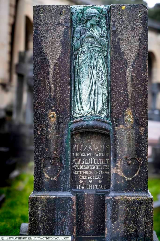 This is a close-up shot of a dark, ornate gravestone in Brompton Cemetery, United Kingdom. The stone features a bronze relief of an angel and an inscription below commemorating Eliza Ann, the beloved wife of Alfred Pettitt, who died in 1896 at the age of 38.