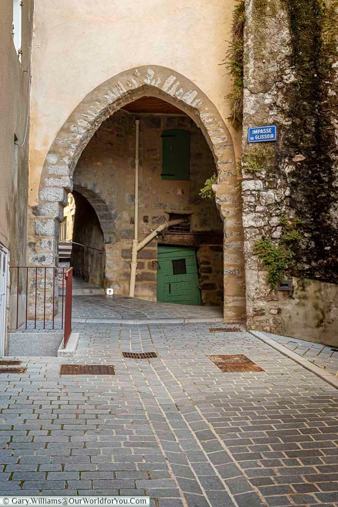 A picturesque, ancient stone archway frames a narrow, cobblestone alley in the historic town of Sisteron, Provence, France, inviting exploration of its hidden passages. A blue street sign reading "Impasse du Glissoir" is visible on the textured stone wall to the right, marking this charming example of secret France waiting to be discovered.