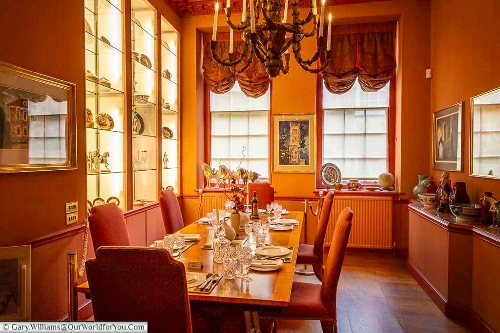 The photograph showcases a beautifully appointed dining room within Arundells House in Salisbury, featuring warm orange walls, a polished wooden floor, and a long dining table set with elegant tableware and surrounded by red upholstered chairs. A grand chandelier hangs above the table, casting a soft glow on the scene, while display cabinets filled with decorative china and artwork adorn the walls.