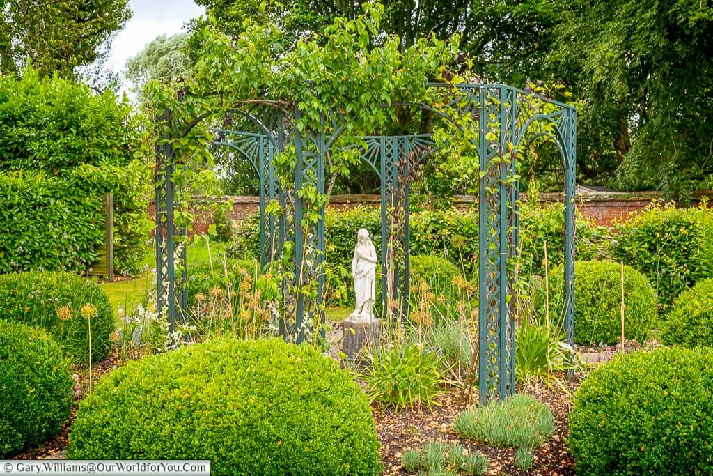 A picturesque garden scene at Arundells house in Salisbury, featuring a classical white statue standing beneath an elegant, green-painted metal pergola entwined with climbing plants. Lush green topiary bushes and a variety of flowering plants fill the foreground and surround the central feature, with a red brick wall and mature trees visible in the background under a cloudy sky.