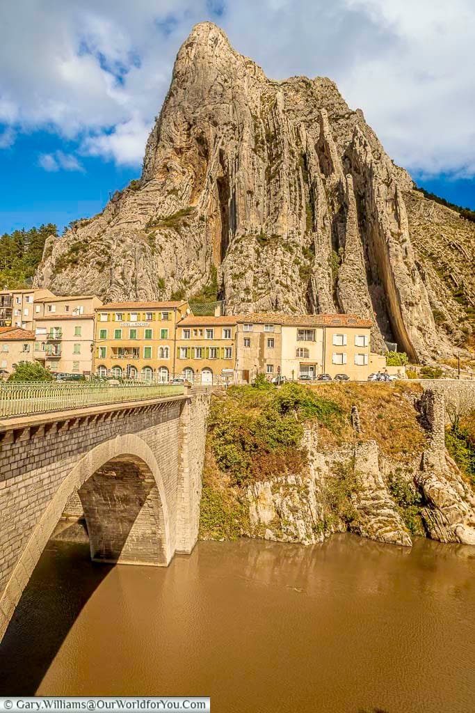 The imposing Rocher de la Baume (Baume Rock) dramatically towers over the historic town of Sisteron in Provence, France, with a classic stone arch bridge crossing the Durance River in the foreground. Colourful buildings huddle at the base of the sheer, textured cliff face, all under a bright blue sky with scattered clouds, creating an iconic French vista.