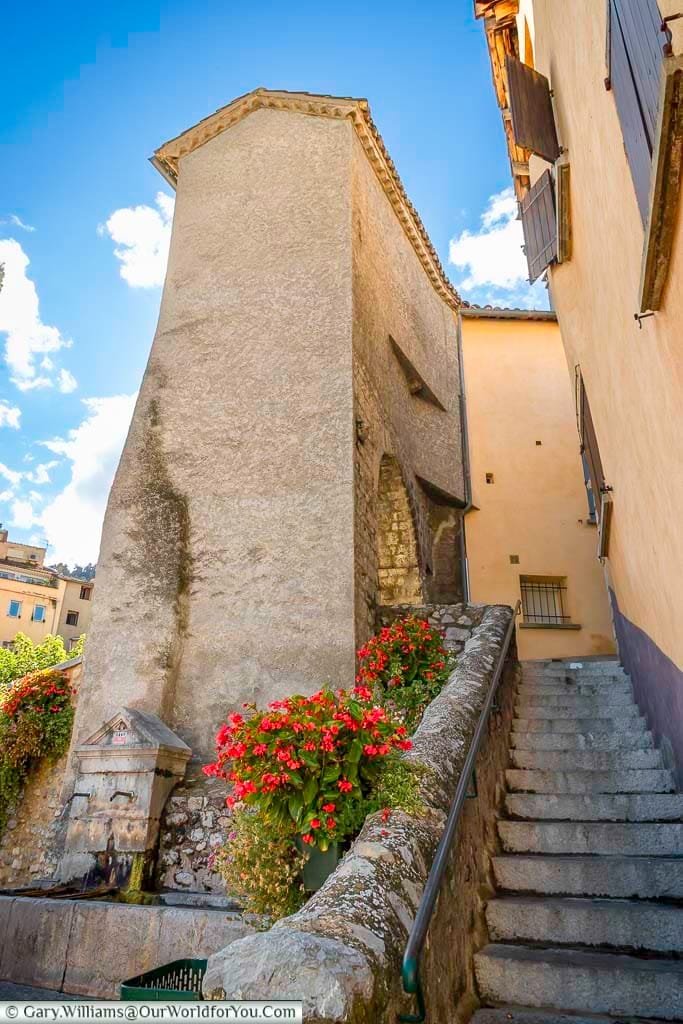 A narrow stone stairway winds up a charming lane in Sisteron, Provence, France, flanked by a historic, light-coloured stone building on one side and vibrant red flowers cascading over a rustic stone wall and fountain on the other. This picturesque hidden corner of France, with its traditional architecture and glimpse of bright blue sky, beckons exploration for those touring the region.