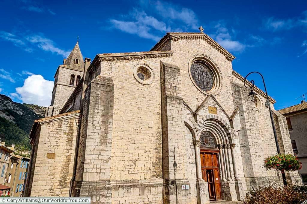 A Romanesque-style stone church with arched doorways and a tall bell tower stands under a bright blue sky in Sisteron, France. The Church of Notre-Dame-des-Pommiers is framed by surrounding buildings and mountain scenery in the background.