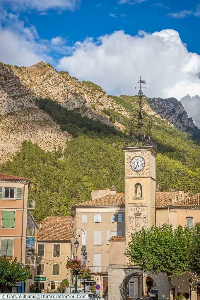 A prominent stone clock tower with an ornate ironwork campanile and weathervane stands tall in Sisteron, France, against a backdrop of a steep, tree-covered mountainside and a bright blue sky with fluffy white clouds. Surrounding the base of the clock tower are traditional European buildings with terracotta tiled roofs and shuttered windows.