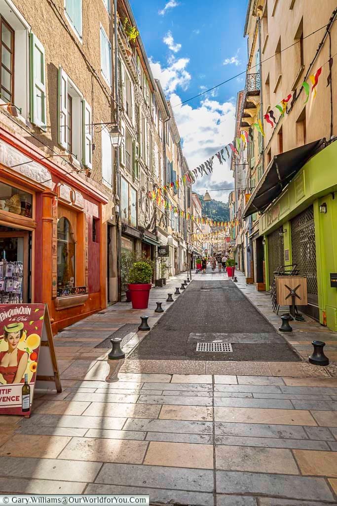 A vibrant, sun-dappled narrow street in Sisteron, Provence, France, is lined with colourful shopfronts painted in hues of orange and lime green, adorned with traditional shutters and festive bunting strung overhead. This charming French street scene, with its patterned paving stones and distant figures, invites exploration and offers a glimpse into the hidden gems of Provence-Alpes-Côte d'Azur.