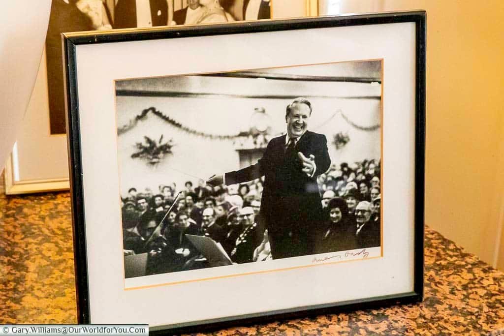 A framed black and white photograph rests on a surface at Arundells House in Salisbury, capturing a smiling ted heath in a suit conducting a large group of people. The image, signed in the bottom right corner, is presented with a white matting and a black frame.