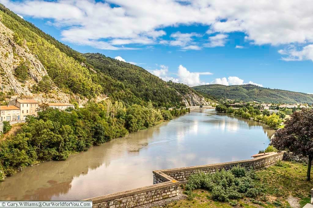 A scenic view of the Durance River near Sisteron in Provence, France, shows its calm waters reflecting the sky as it winds through lush, tree-covered hills under a partly cloudy blue sky. In the foreground, a stone wall and some vegetation frame the picturesque French countryside, with a glimpse of Sisteron town nestled in the distance, making it an inviting scene for those touring France.