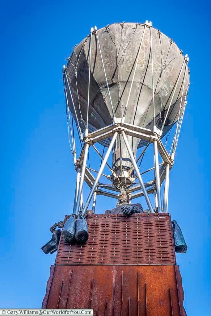 This image depicts the Balloon of Friendship sculpture by Dany Tulkens in Leuven, Flanders, Belgium, set against a clear blue sky. The sculpture features a large, gray balloon-like structure atop a geometric metal frame and a red, textured base, with small, dark objects hanging from the basket.