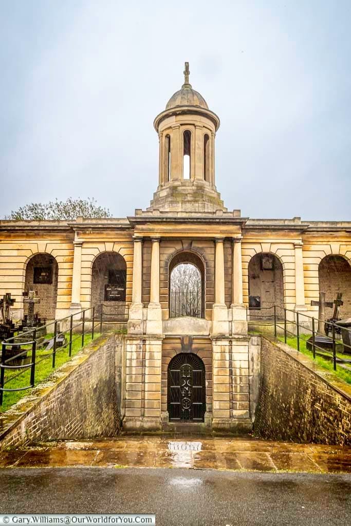 This eye-level shot shows a grand, classical-style mausoleum at Brompton Cemetery in the United Kingdom under a cloudy sky. The structure features arched alcoves, a central domed tower, and a gated entrance leading down a short flight of steps, surrounded by weathered stone and some greenery.