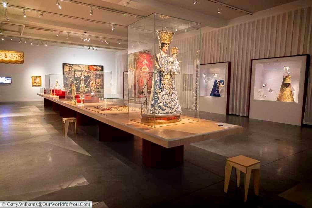 This interior shot captures a brightly lit exhibition space at M - Museum Leuven in Flanders, Belgium. A long, light-colored display table runs through the center, showcasing various religious artifacts under glass cases, while additional exhibits are visible against the white walls in the background.