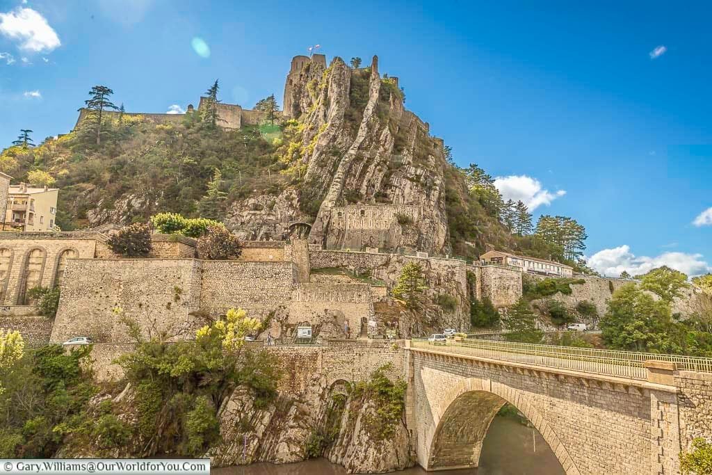 The majestic Sisteron Citadel in Provence, France, stands perched atop a dramatic rocky cliff, overlooking the historic town and the Durance River flowing below a classic stone arch bridge. This iconic French landmark, with its ancient fortifications clearly visible against a bright blue sky dotted with clouds, offers a breathtaking view of hidden Provence.