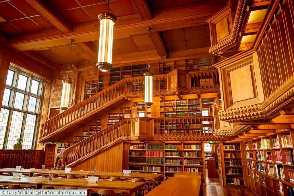 This image captures the grand interior of the main hall in the University Library of Leuven, Flanders, Belgium. The hall features towering wooden bookshelves, ornate staircases, and rows of tables for study, all illuminated by hanging lamps.