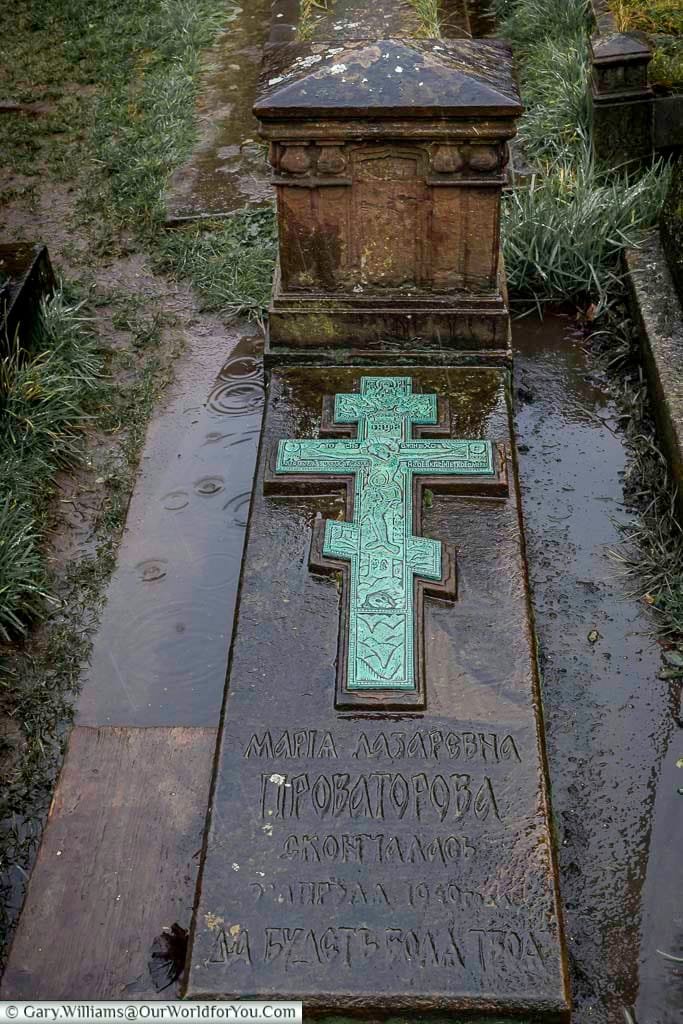 This image shows a close-up of the grave of Maria Lazarevna Glukhareff Provetoroff in Brompton Cemetery. The dark stone features a prominent, ornate green cross and inscriptions in Cyrillic script.