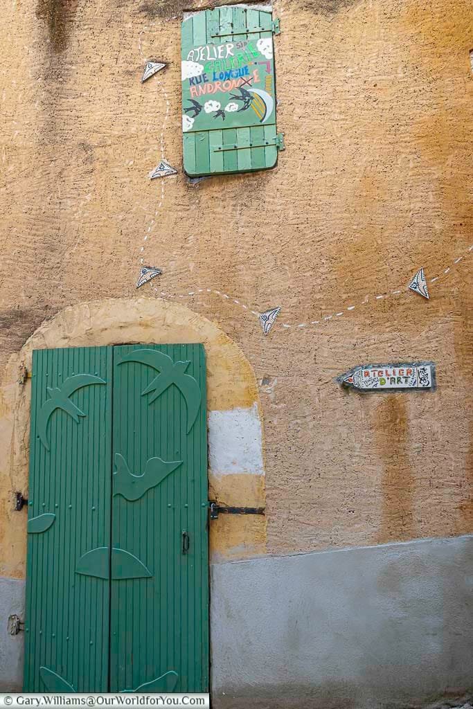 A rustic, textured ochre wall on a medieval street in Sisteron, Provence, France, features a vibrant green wooden door adorned with charming bird silhouette carvings and a matching green shutter above, painted with "Atelier" and artistic designs. Small, decorative arrow-like tiles create a whimsical trail across the wall towards another "Atelier d'Art" sign, highlighting the artistic French charm found when exploring Sisteron.